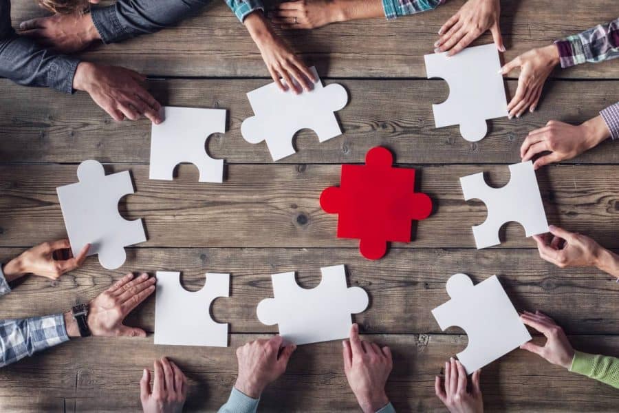 Birdseye view of multiple hands touching large, white puzzle pieces while a red puzzle piece sits in the middle