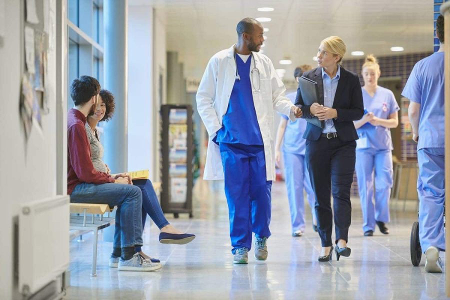 A doctor and hospital administrator walk down a hospital hallway by nurses and patients