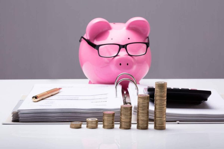 Piggy bank wearing glasses looking at an open binder with paper and a calculator with stacks of coins in front