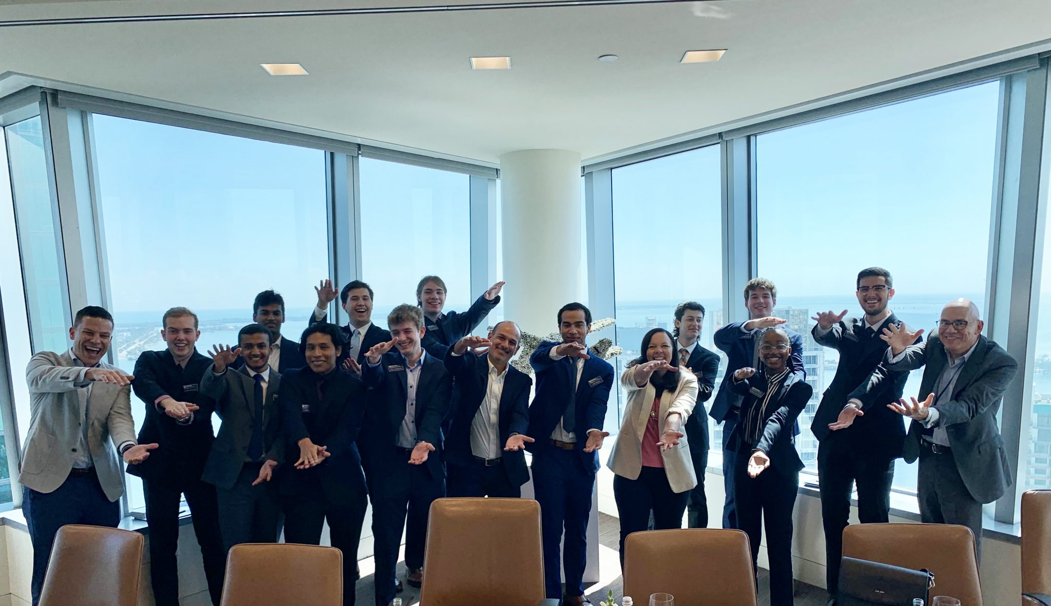 A group of 15 students stand in front of a large window while doing the Gator Chomp.