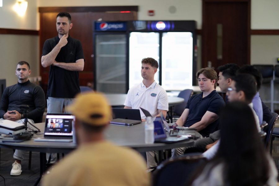 A man stands near a table as others are seated with laptops open.