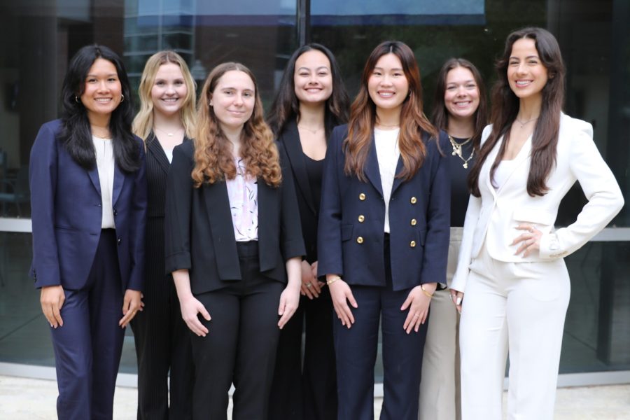 A group of seven students in business attire smiling in front of Hough Hall.