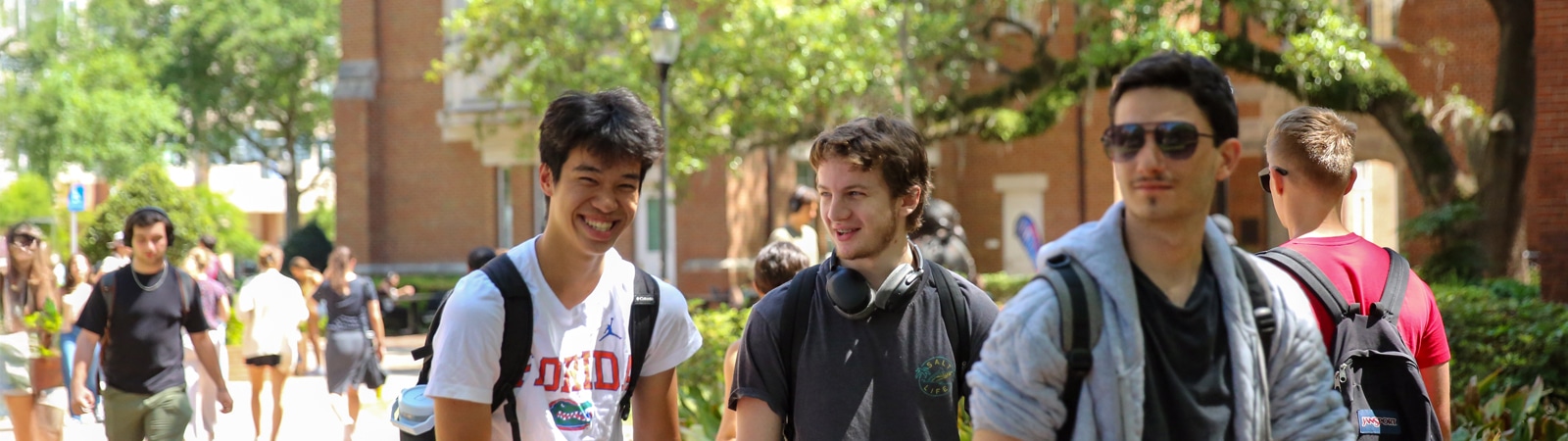 Three casually dressed students walk and talk on the sunlit college of business campus surrounded by trees and brick buildings.