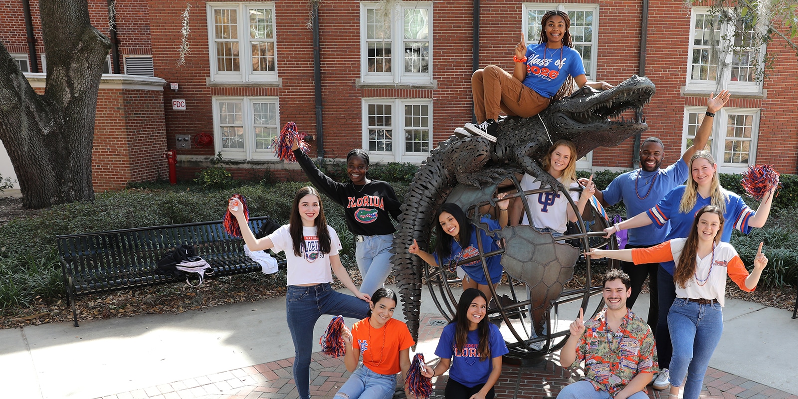 Recent University of Florida Warrington College of Business alumni hold up fingers or wave pom-poms while standing around the campus Gator statue