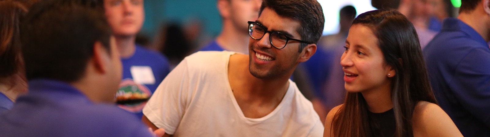 A group of young adults smiling and engaging in conversation at a social event. The atmosphere is lively and informal, with people wearing casual clothes and mingling in the background.