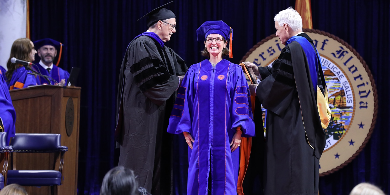 In a cap and gown, a UF Warrington DBA student smiles on stage while being presented with their diploma
