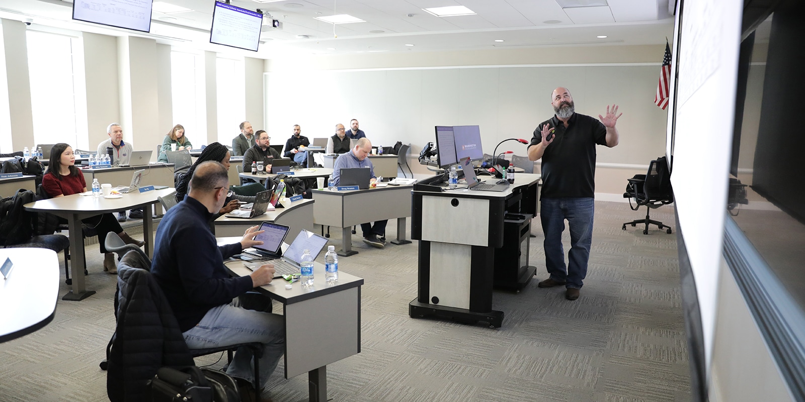 Warrington DBA students sitting at individual desks in a classroom listen to a professor's presentation using the screen behind