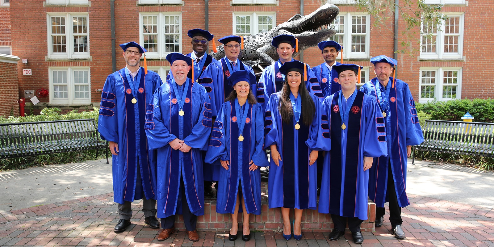 A group of DBA students in caps and gowns stand in front of the University of Florida's Gator statue