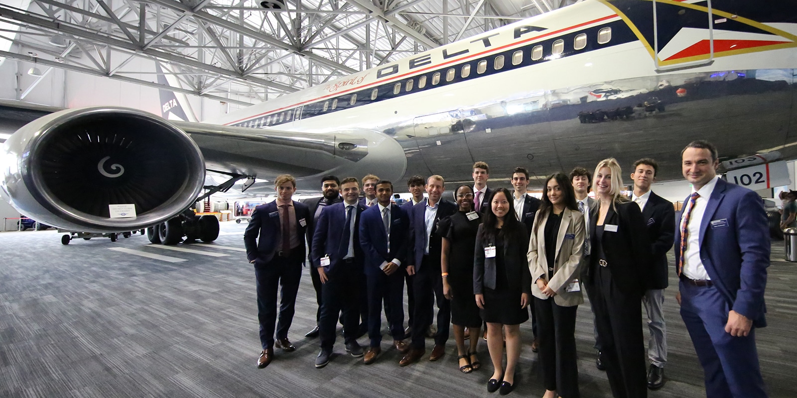 A group of University of Florida Warrington College of Business students in suits stand in front of a Delta airplane inside a hangar