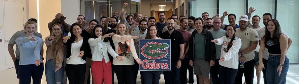 A group of students from UF Warrington's South Florida Weekend Part-Time MBA program make Gator arms while standing inside campus and holding a sign with the school's mascot