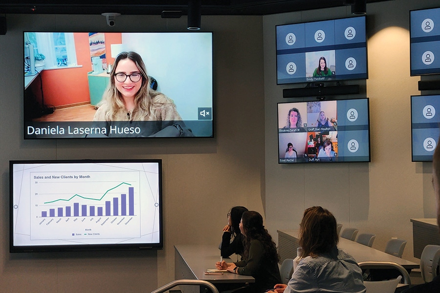 Students participating in a video conference at the University of Florida Warrington College of Business, with presentation slides and multiple screens visible.