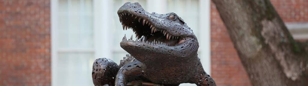 A statue of the University of Florida's alligator mascot, with its mouth open, made of metal, positioned against a brick building background.