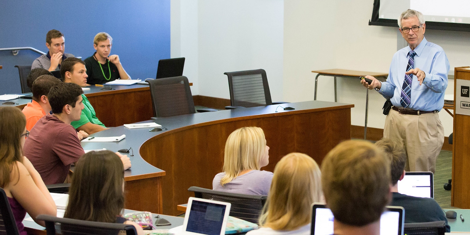 A professor lecturing at the University of Florida Warrington College of Business, with attentive students seated in a classroom with laptops and notebooks.