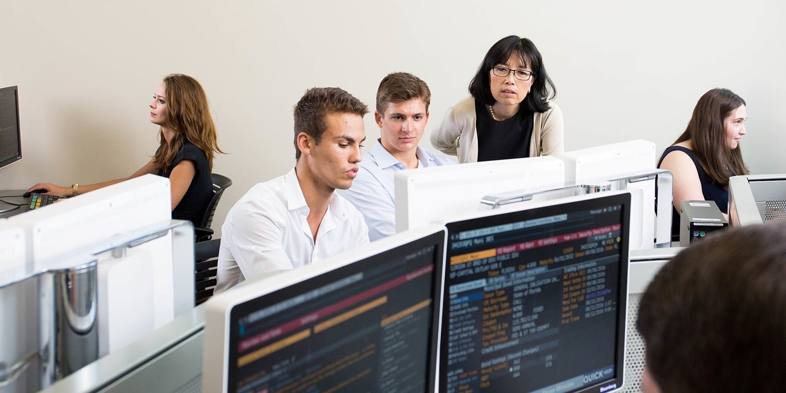 University of Florida Warrington College of Business students work at computer stations using data and AI tools, guided by a mentor, focusing on multiple screens displaying information.
