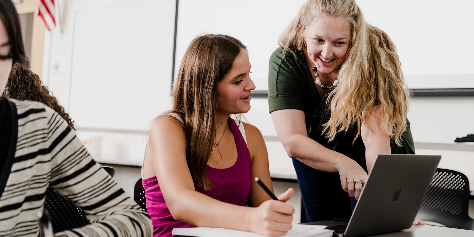A classroom at the University of Florida Warrington College of Business, where a student receives guidance from a teacher while working on a laptop, both smiling and engaged.
