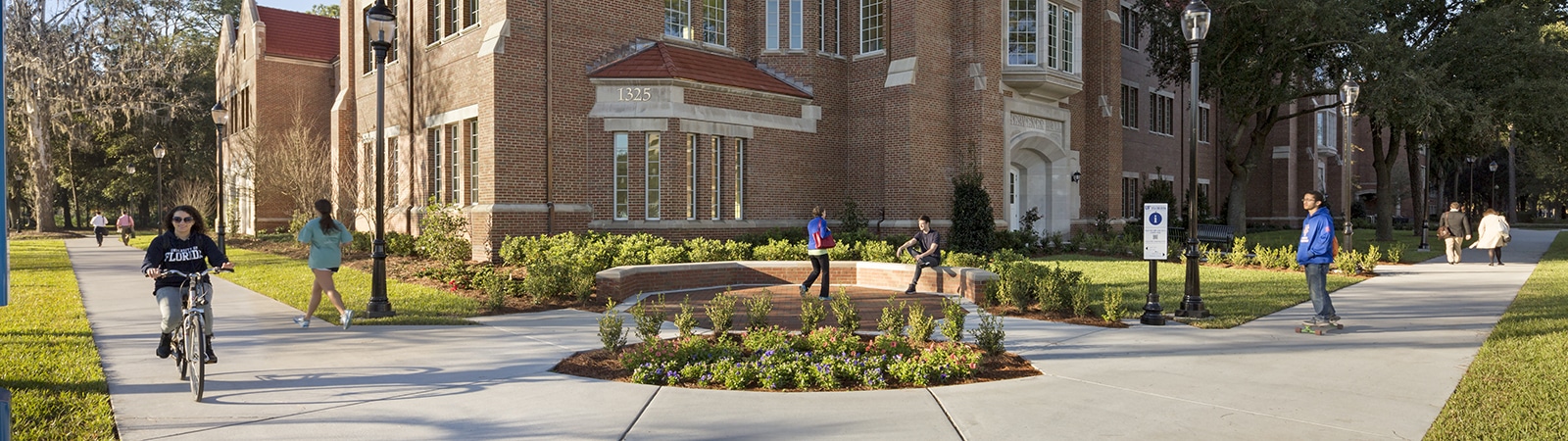 The University of Florida Warrington College of Business building, with people walking and biking on the paths surrounded by landscaped gardens.