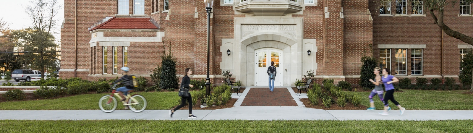 Students jog, bike, and walk in front of Heavener Hall at the University of Florida, a classic red-brick building that houses the Warrington College of Business.