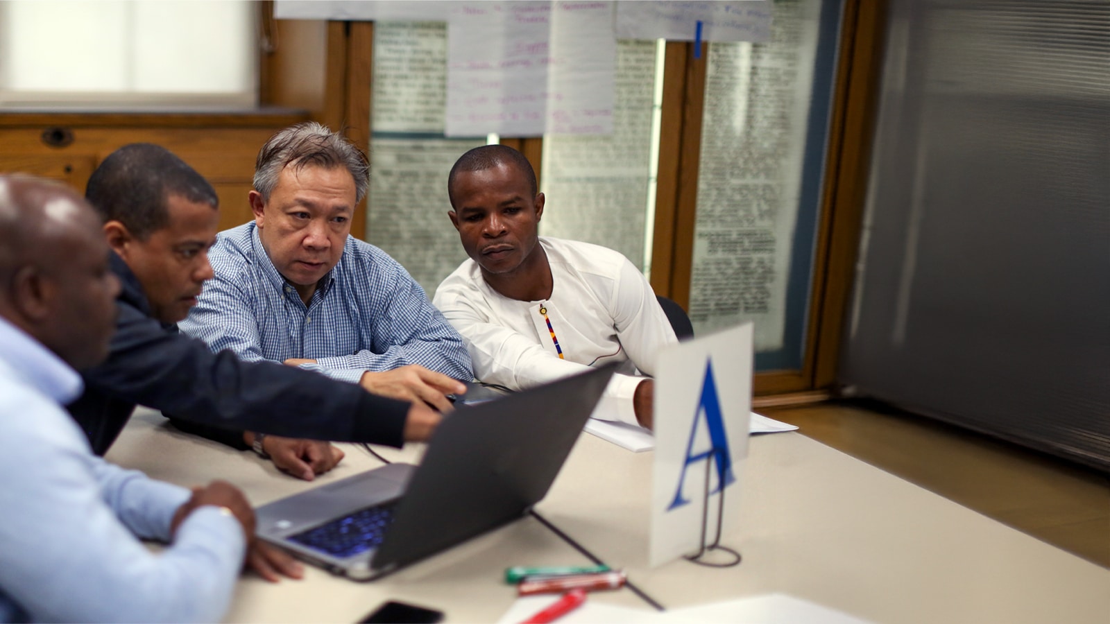 Four men sit at a table focused on a laptop, collaborating during a workshop.