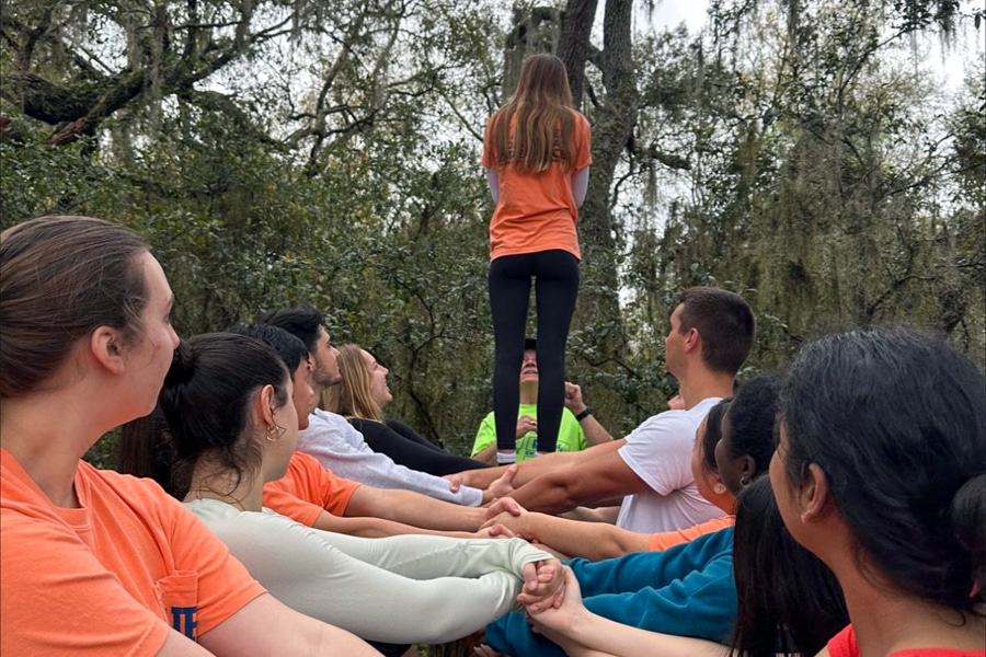 A group of students outdoors participate in a trust-building activity, supporting a teammate standing on their joined hands in a wooded setting.