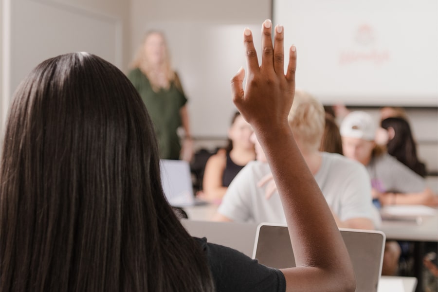 A student with long dark hair raises her hand in a classroom while a faculty member stands in front, with other students listening attentively.