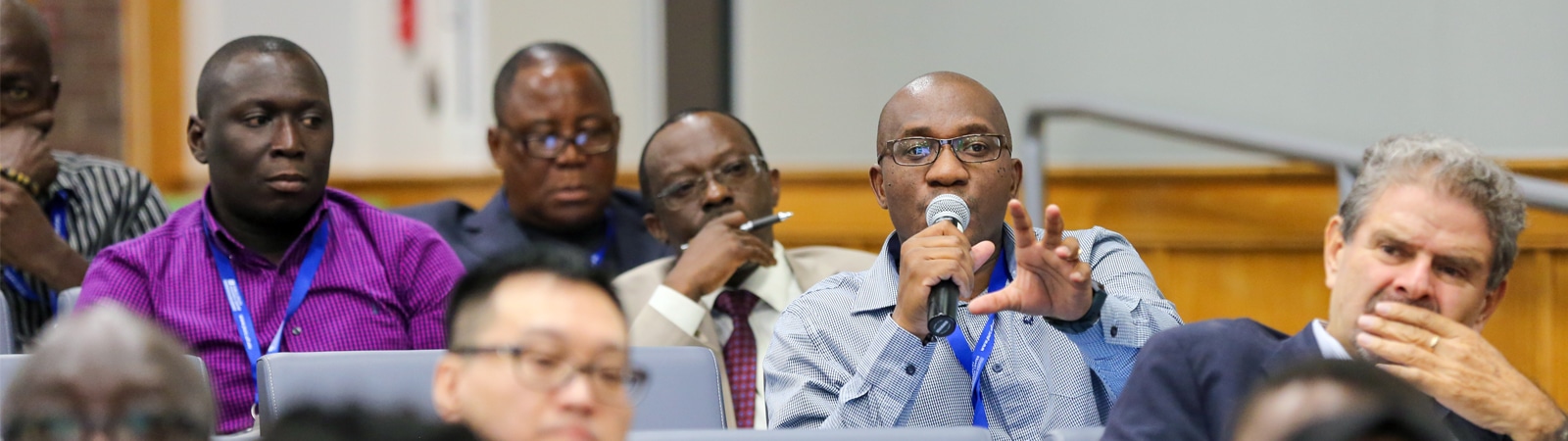 A group of participants in a conference auditorium, with one man speaking into a microphone, engaging in discussion during a PURC training session.