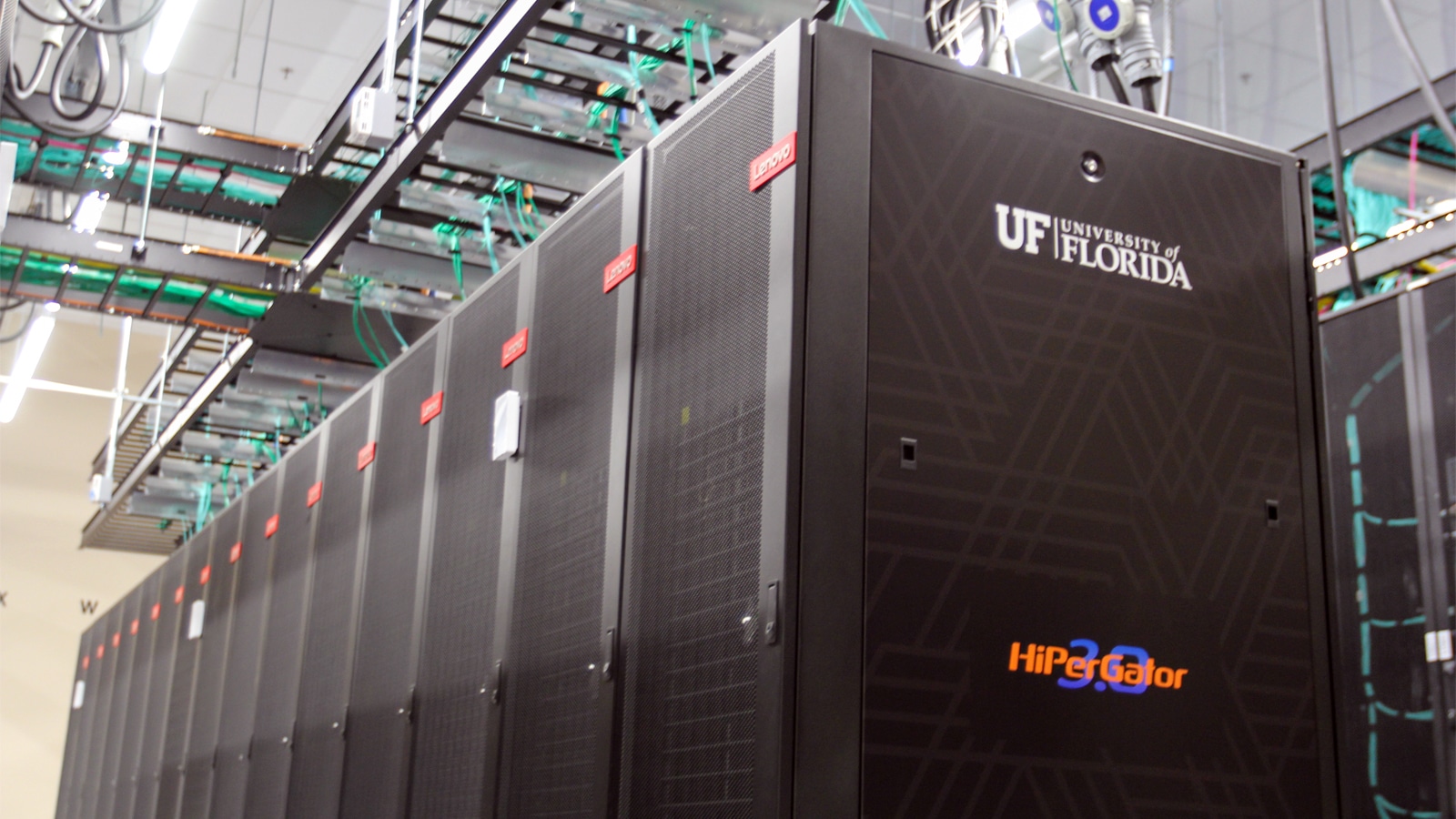 A row of black server cabinets labeled “HiPerGator 3.0” and branded with the University of Florida logo, situated in a technology center with overhead wiring and lighting.