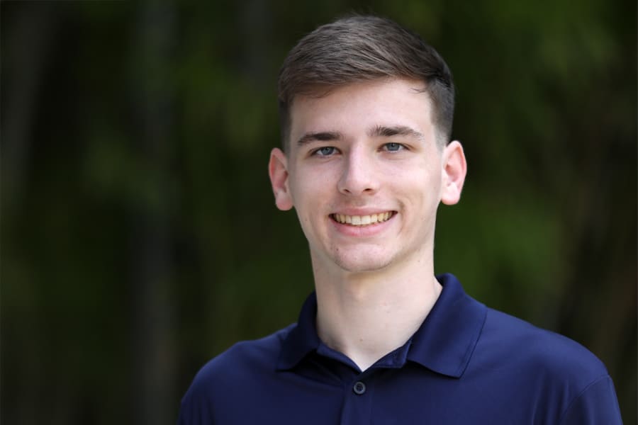 A young man with short brown hair smiles at the camera, dressed in a navy blue Leadership Development Program polo shirt, standing outdoors with greenery in the background.