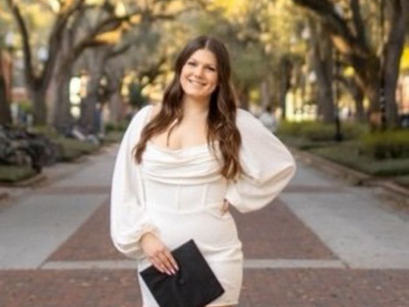 A young woman with long, wavy brown hair and a bright smile poses confidently in a white dress, holding a black clutch while standing on a picturesque, tree-lined brick walkway.