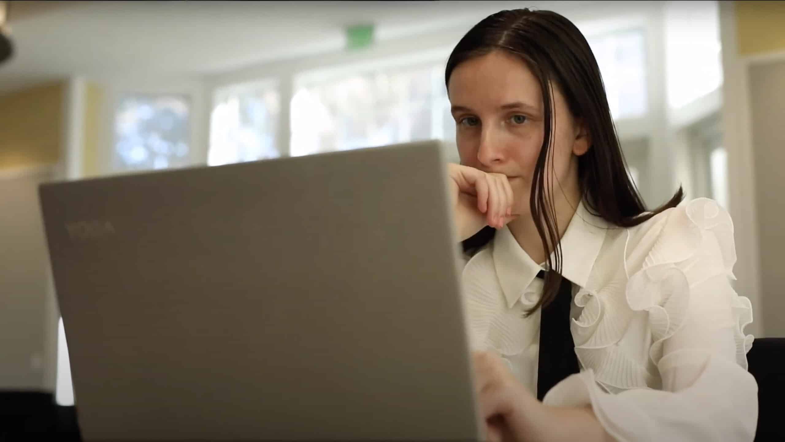 A focused business student, dressed in a white ruffled blouse and black tie, intently works on a laptop in a modern workspace.