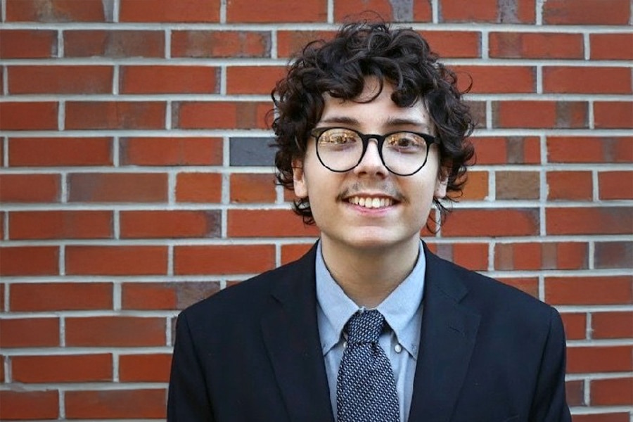 A young man with curly hair, glasses, and a mustache, wearing a dark suit, light blue shirt, and patterned tie, smiling warmly against a red brick wall background.