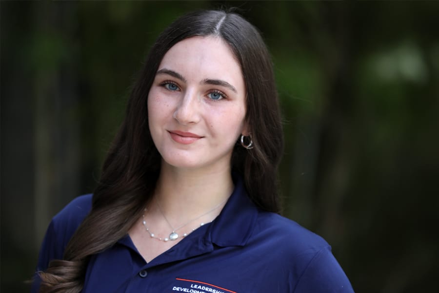 A young woman with long, dark brown hair and blue eyes smiles softly at the camera, wearing a navy blue Leadership Development Program polo shirt, with a natural outdoor background.