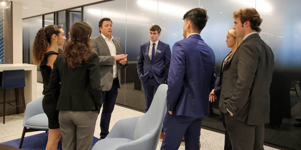 During an internship placement, a group of BSBA in Information Systems students from UF Warrington wearing suits listen to a corporate sponsor discuss company culture
