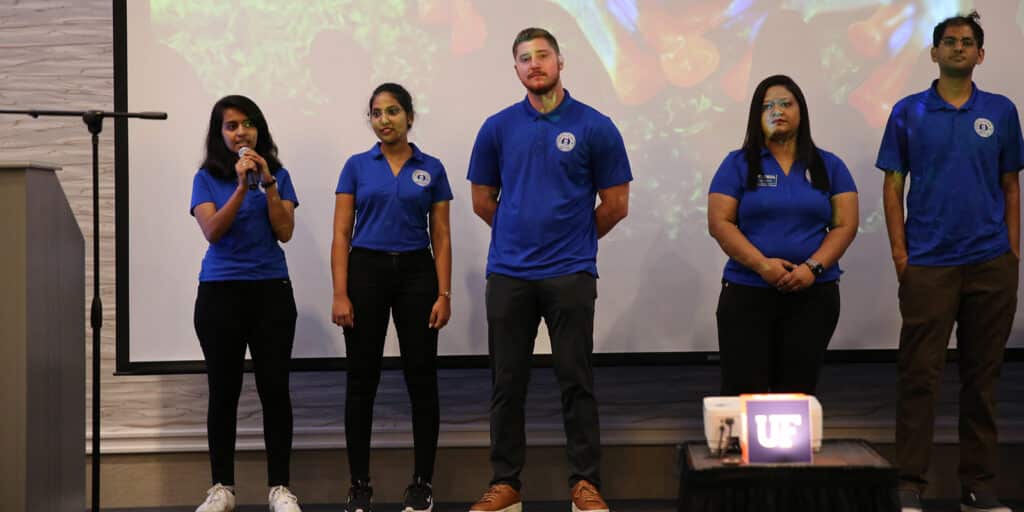 A group of UF Warrington BSBA in Information Systems students in blue shirts stand in front of a screen during a presentation