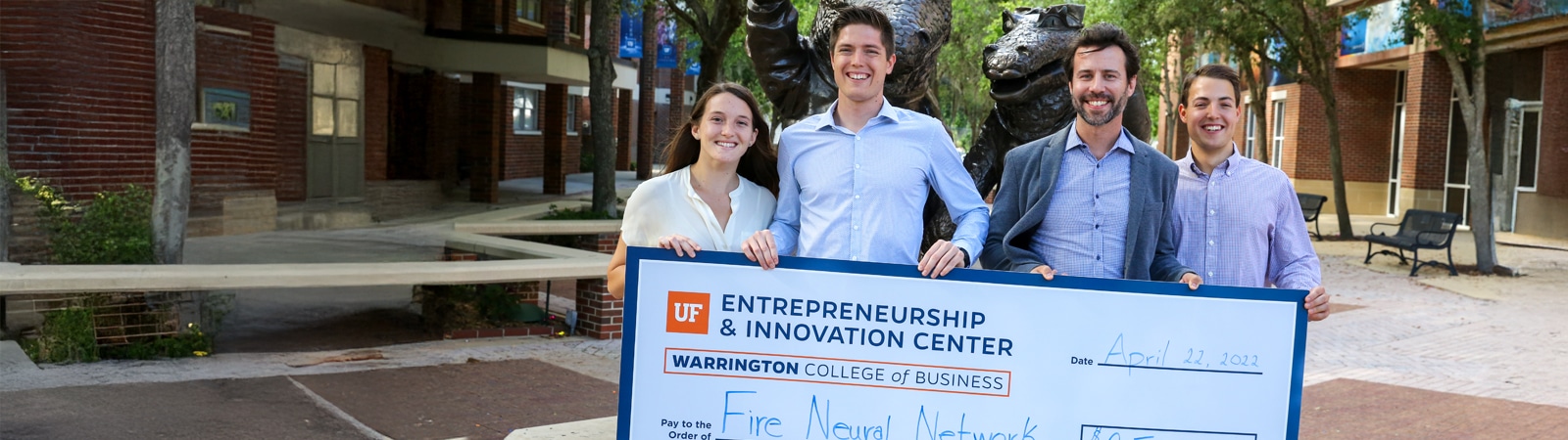 Four smiling young entrepreneurs standing outdoors holding an oversized ceremonial check from the UF Entrepreneurship & Innovation Center, awarded to “Fire Neural Network.”