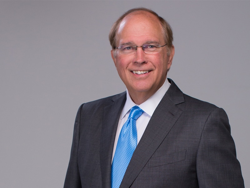 Bill Heavener wearing a grey suit and blue tie, smiling in front of a light grey background.