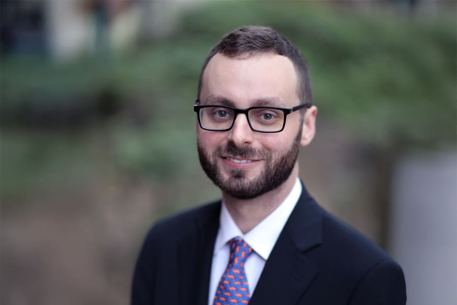 Portrait of Brooks Riggeal, a young man with glasses and a trimmed beard, wearing a dark suit, white shirt, and an orange patterned tie, smiling against a blurred natural background.