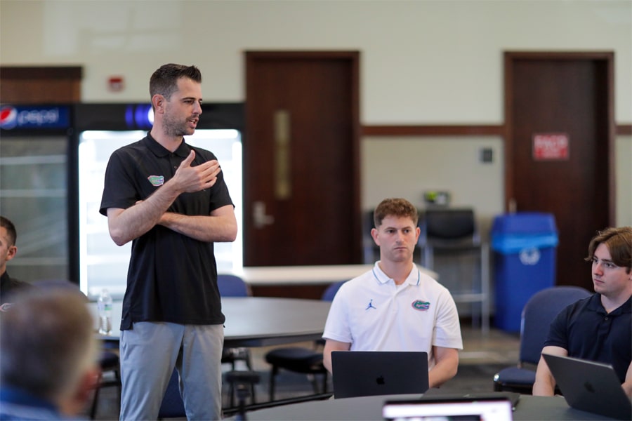Todd Golden stands and gestures as he speaks to a group of students seated around a table, some with laptops open, in a classroom setting. The students listen attentively.