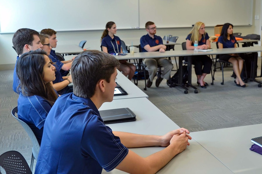 A group of students wearing matching navy blue polos sit attentively in a classroom arranged in a U-shaped table layout.