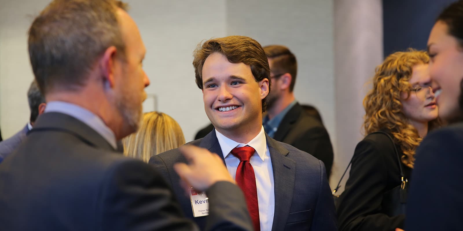 Business people mingling with the focus on a smiling young man in a suit and tie.