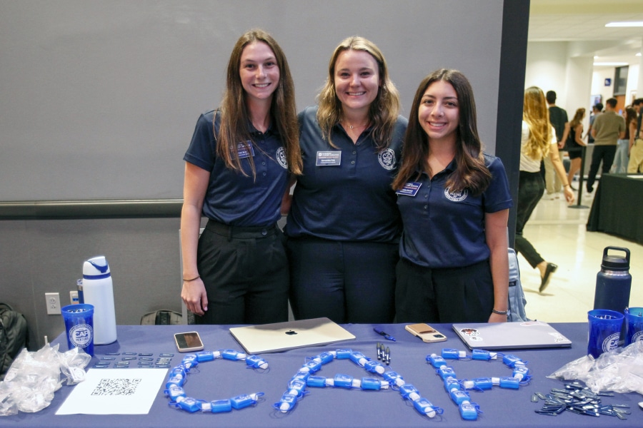 Three female student mentors in matching navy blue polo shirts stand smiling behind a table at a career event, with promotional items arranged to spell “CAP.”