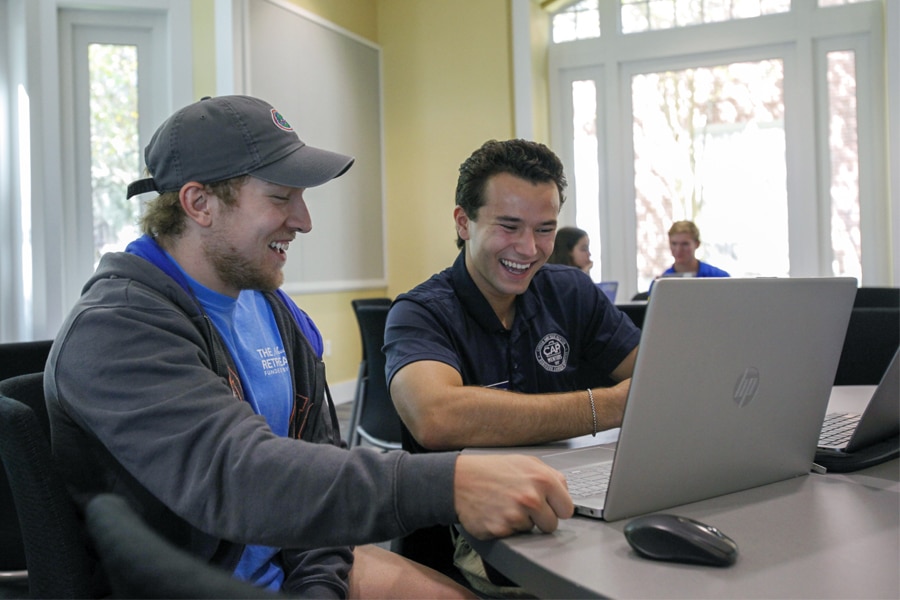 Two University of Florida graduate students sit side by side at a table, smiling and looking at a laptop screen in a bright, open room with large windows and other students in the background.