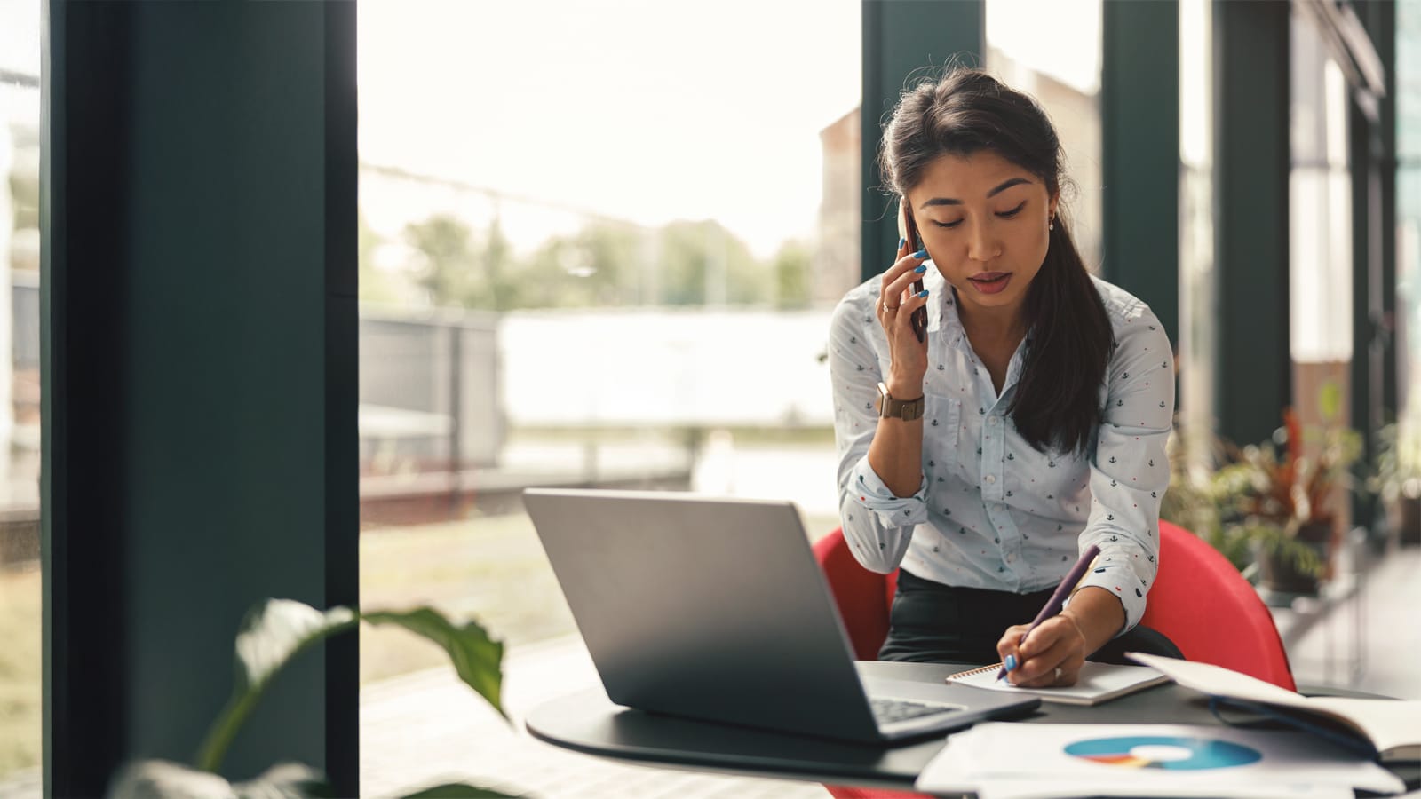 A woman sits at a table in a modern office, talking on her phone while writing notes in a notebook, with a laptop and documents spread out in front of her.