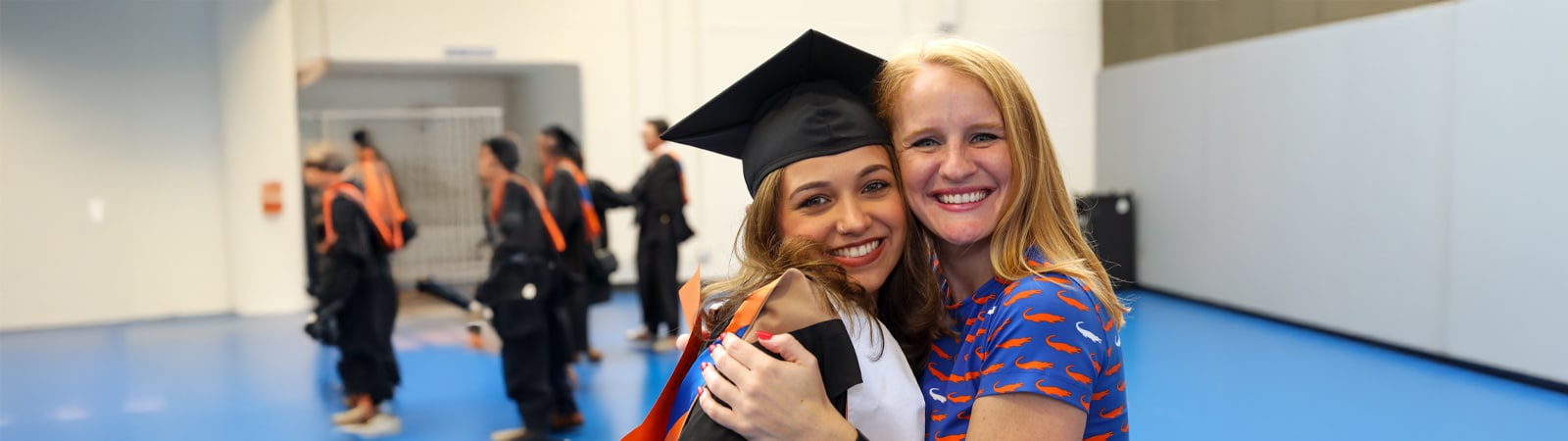 A graduate in cap and gown embraces a smiling supporter wearing a Gators‑print shirt in an indoor staging area before Commencement.