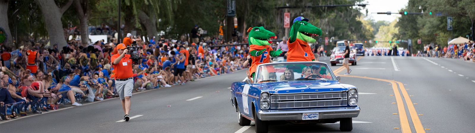 UF mascots Albert and Alberta ride in a blue convertible during a parade, waving to a crowd lining the street.