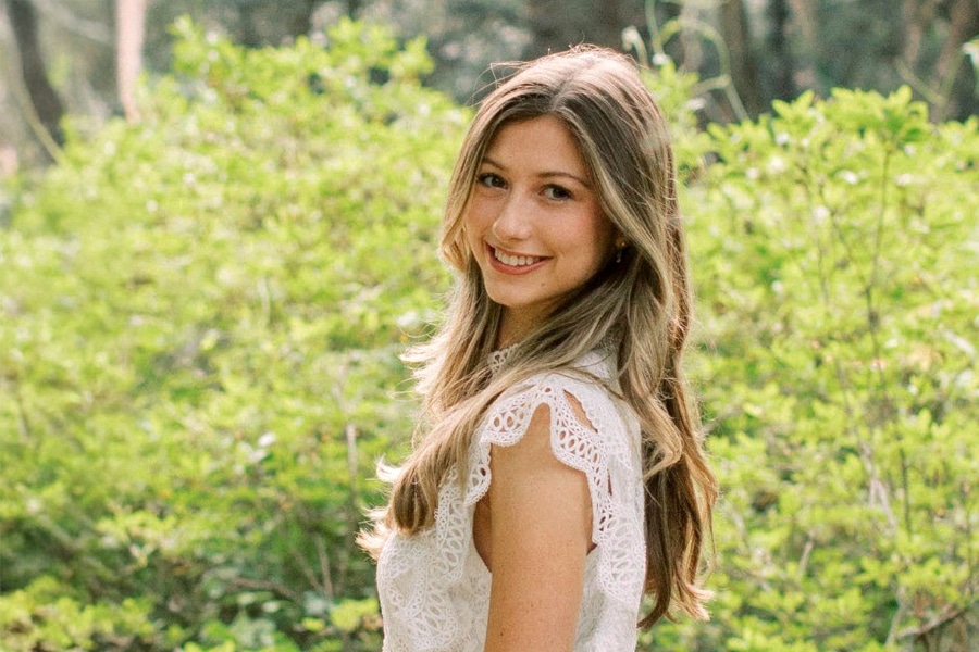 A young woman with long, light brown hair smiles as she looks over her shoulder, wearing a white lace top and standing outdoors in front of lush green foliage.