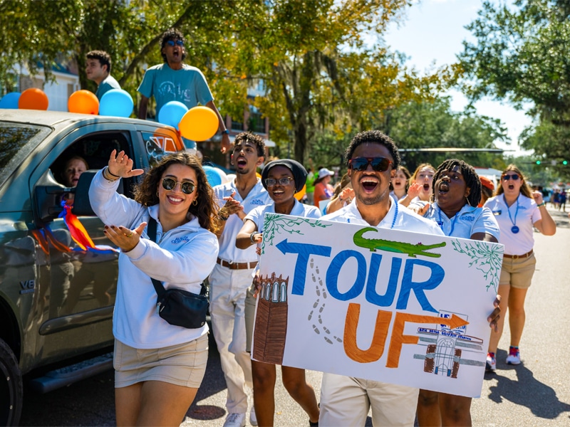 A group of smiling and cheering UF students in white shirts and khaki shorts hold a colorful hand-painted sign that reads “Tour UF” as they walk in a sunny parade. Blue and orange balloons are attached to a car behind them.