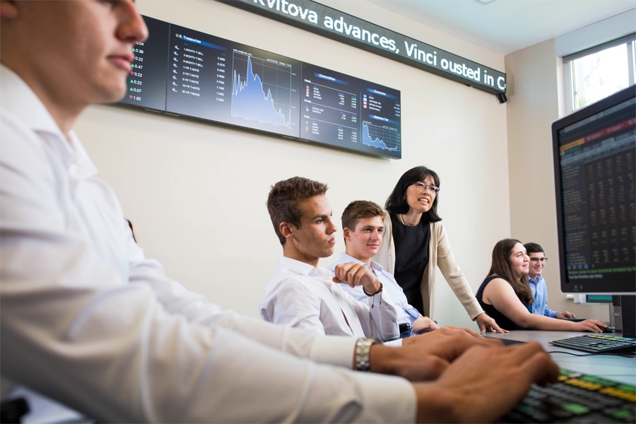 Finance students work at computer terminals displaying financial data, with an instructor guiding them in a modern classroom equipped with digital market tickers and financial charts on the wall.