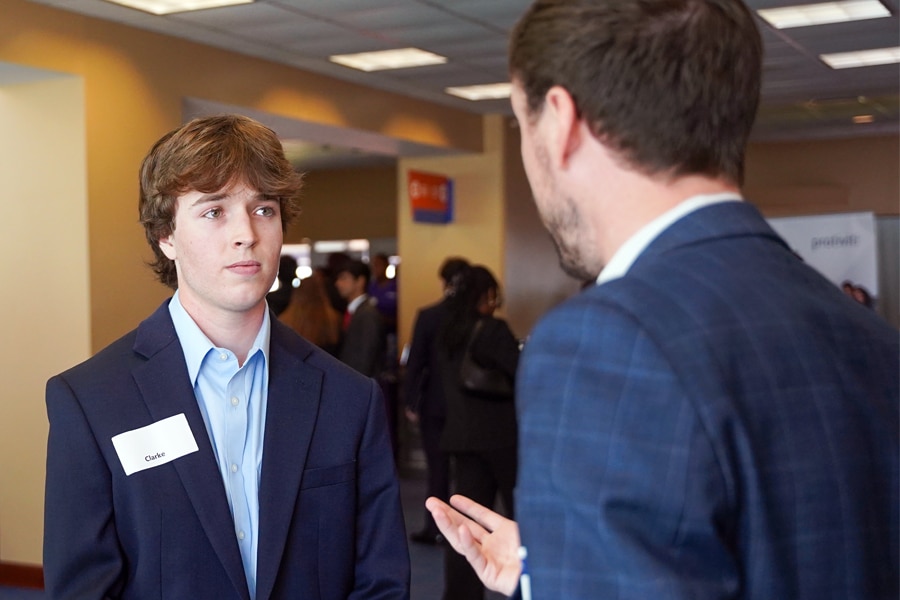 A University of Florida graduate student wearing a name tag labeled "Clarke" listens attentively while a professional coach speaks to him at a career event with other attendees in the background.
