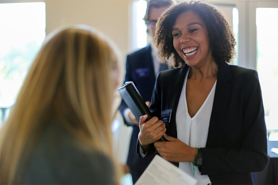 A smiling young woman with curly hair, dressed in business attire, holds a portfolio while engaging in conversation with another person at a career fair.