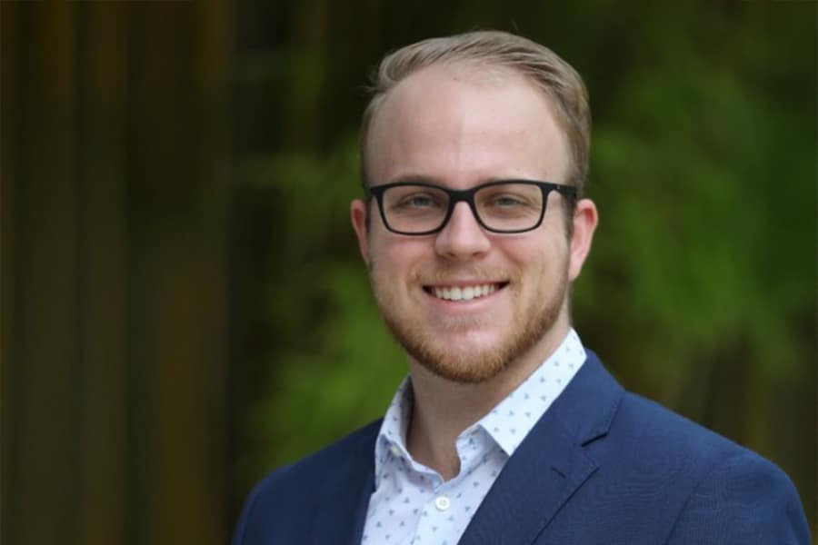 A young man with short blond hair, a trimmed beard, and glasses smiles confidently while wearing a blue suit jacket and patterned white shirt, standing outdoors.